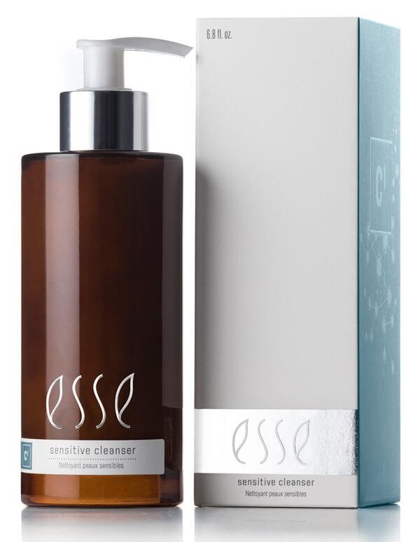 A bottle labeled "Esse Sensitive Cleanser" with a pump dispenser and brown in color is positioned next to a silver and white box that features the same branding. The bottle's label showcases white text, while the box has a metallic strip near the bottom providing product information.